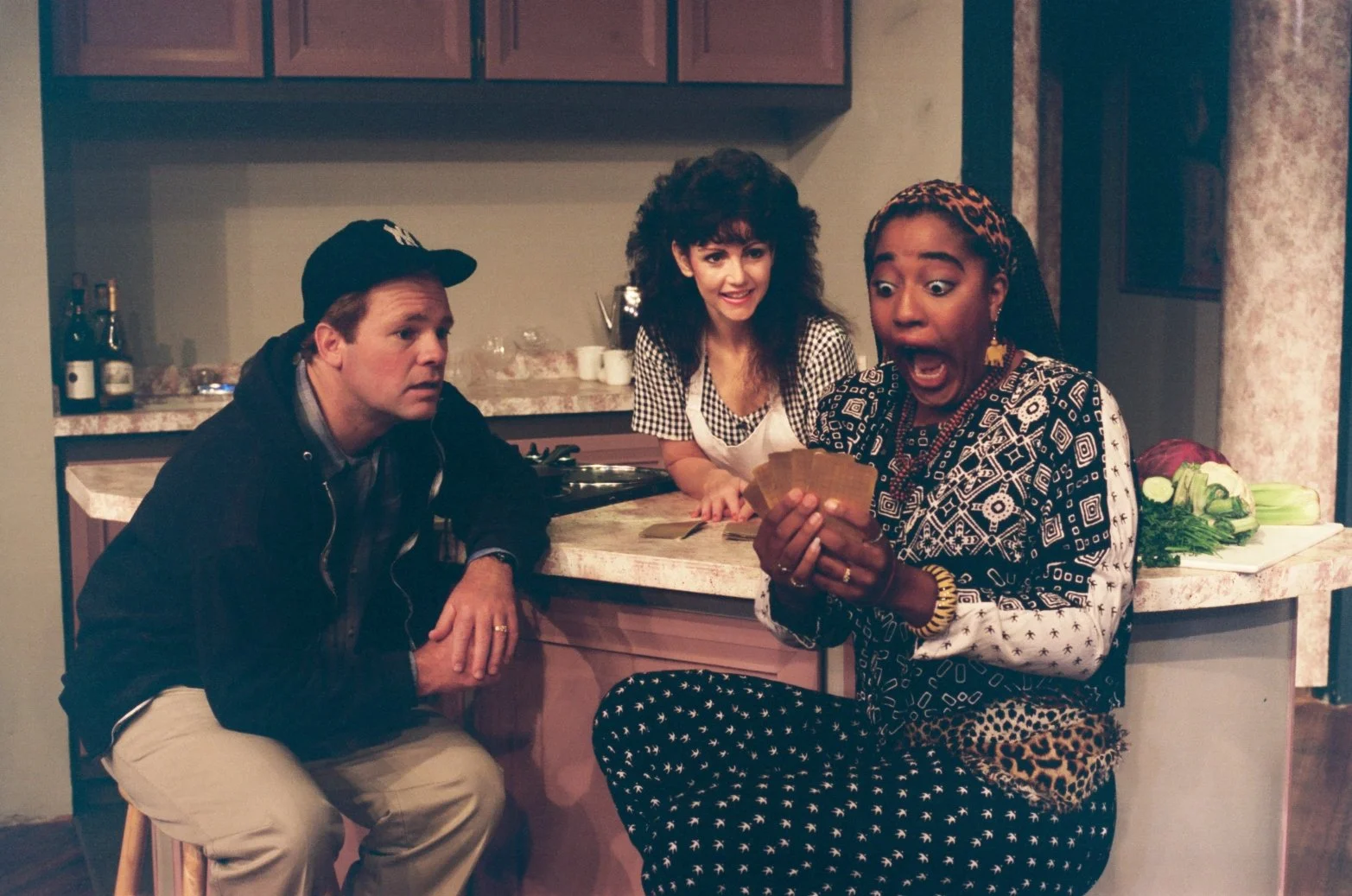 Three people in a kitchen, one wearing a cap, another in an apron, and the third reacting dramatically while holding cards. Bottles are on the countertop.