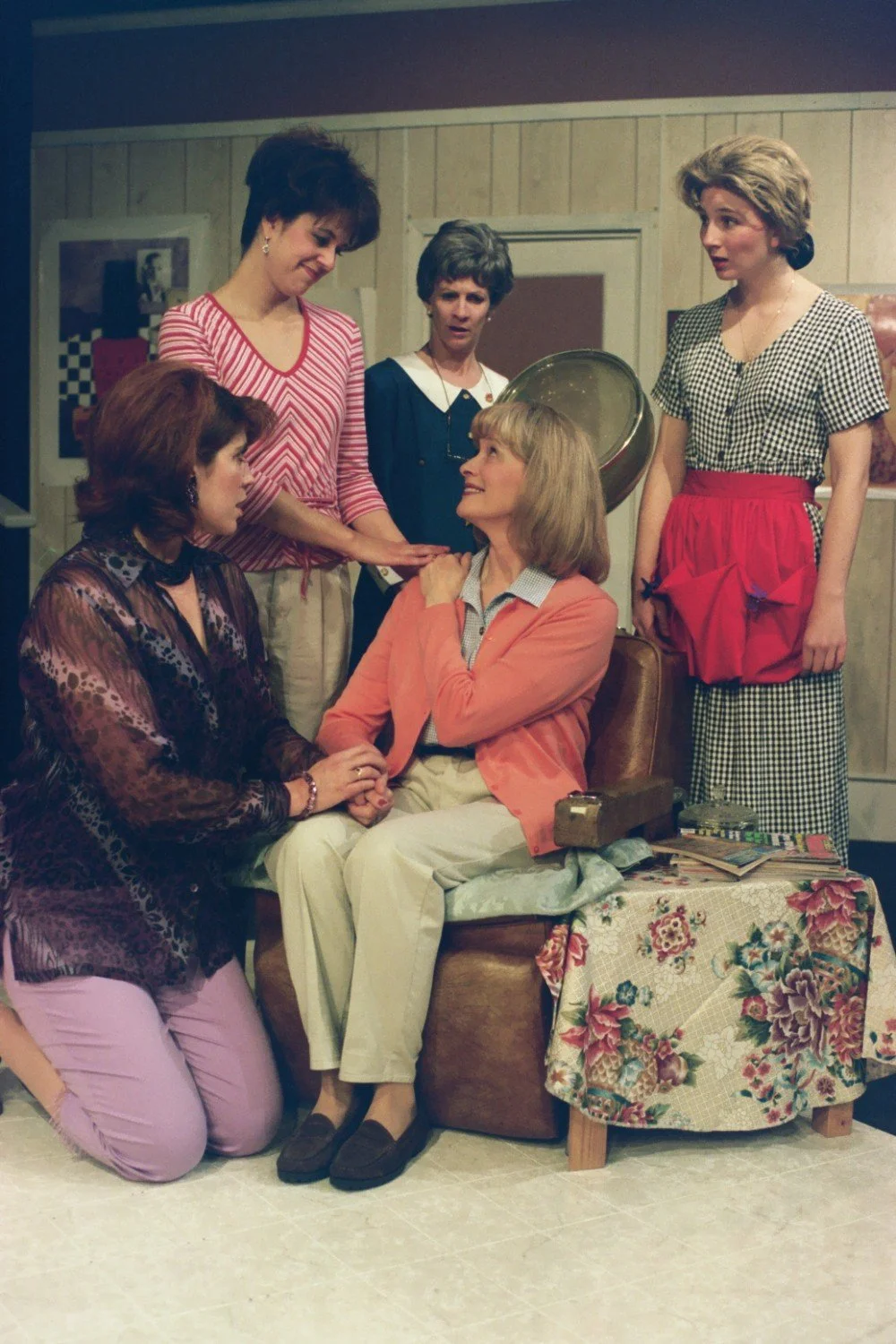 Five women in a theatrical scene, with one seated and the others standing around her. The scene appears to be a drama or play set in a domestic setting.