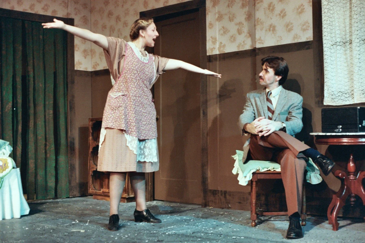 Two actors performing a stage play. The woman is wearing a patterned apron and gesturing with outstretched arms, while the seated man in a suit observes her. The setting includes vintage-style furniture and wallpaper.