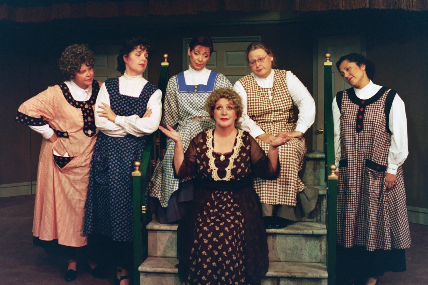 Six women in vintage-style dresses posing on a staircase, with one woman sitting and gesturing with her hands.