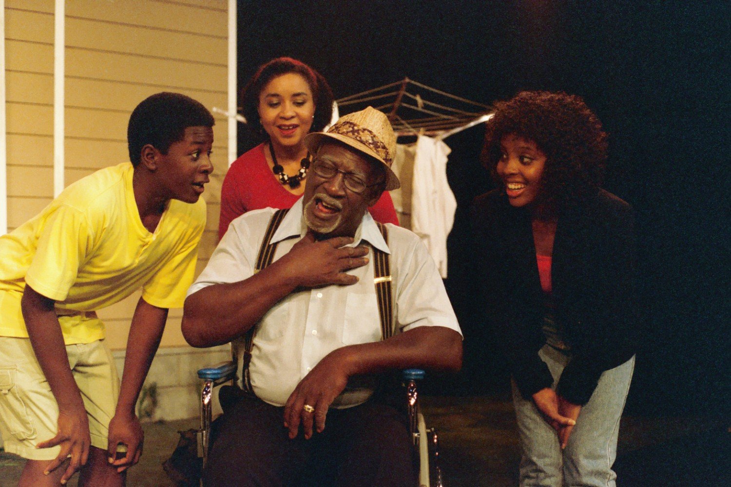 A group of four people, including a man in a wheelchair wearing a hat, are joyfully interacting. They are in an outdoor setting with laundry drying in the background. The group includes a young boy in yellow, a woman in red, and a woman with curly ha