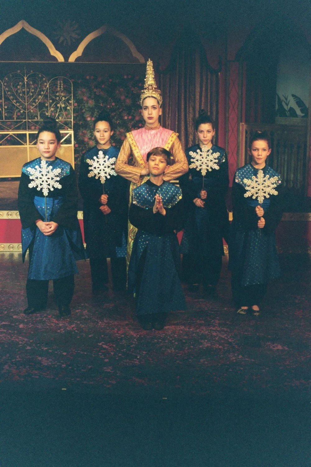 Children in blue costumes holding snowflake props with a person in traditional attire on stage