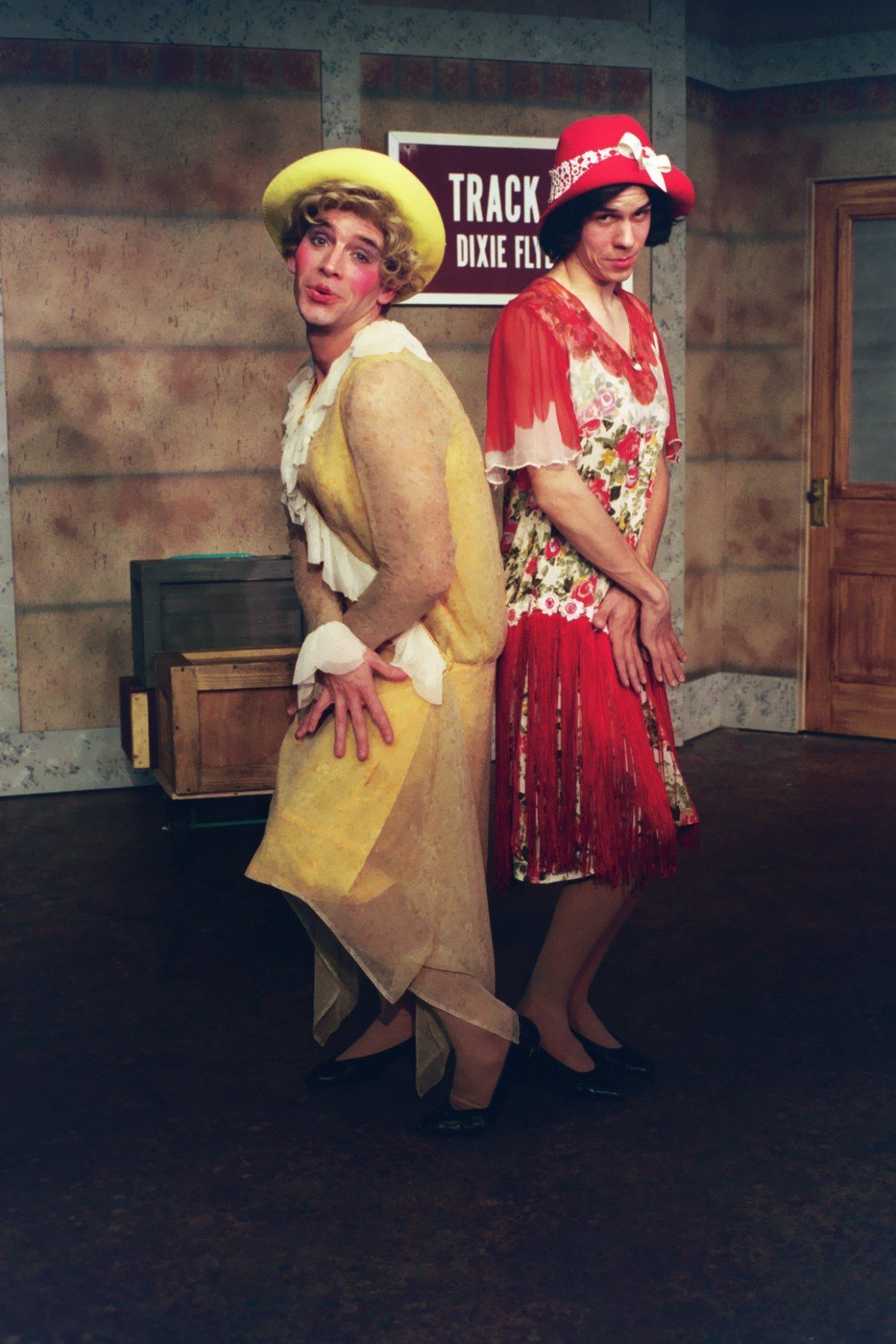 Two people in colorful, vintage-style costumes with hats, standing in a theatrical setting.