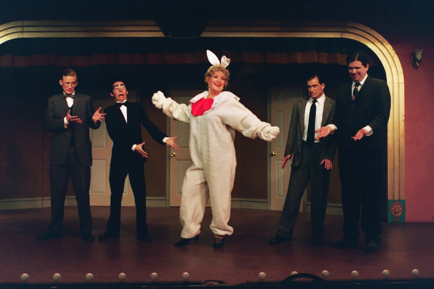 A person in a bunny costume on stage with four men in suits reacting comically.