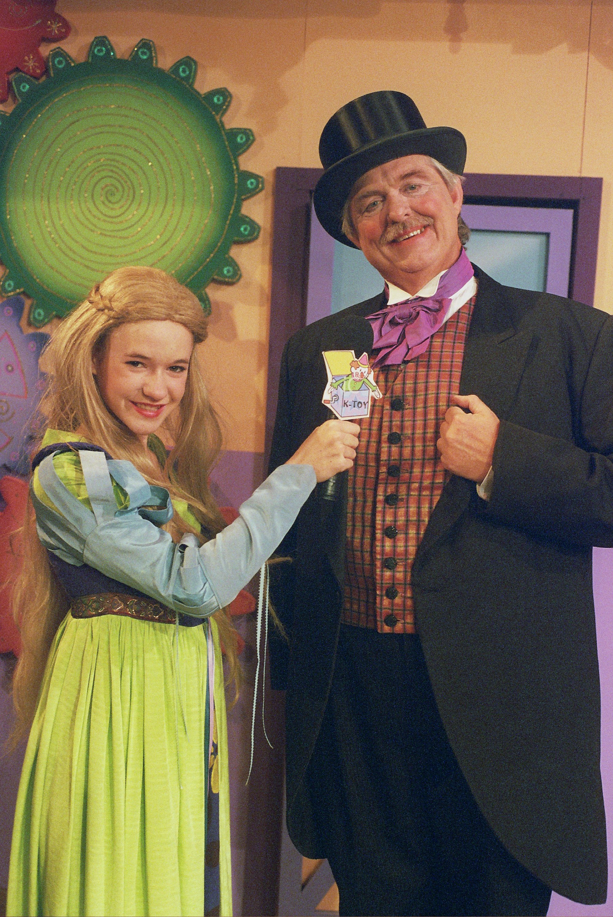 A young woman in a fairy tale costume holding a microphone labeled "K-Toy" and interviewing a man dressed in a top hat and vintage suit with a bow tie. The background features colorful, whimsical decor.
