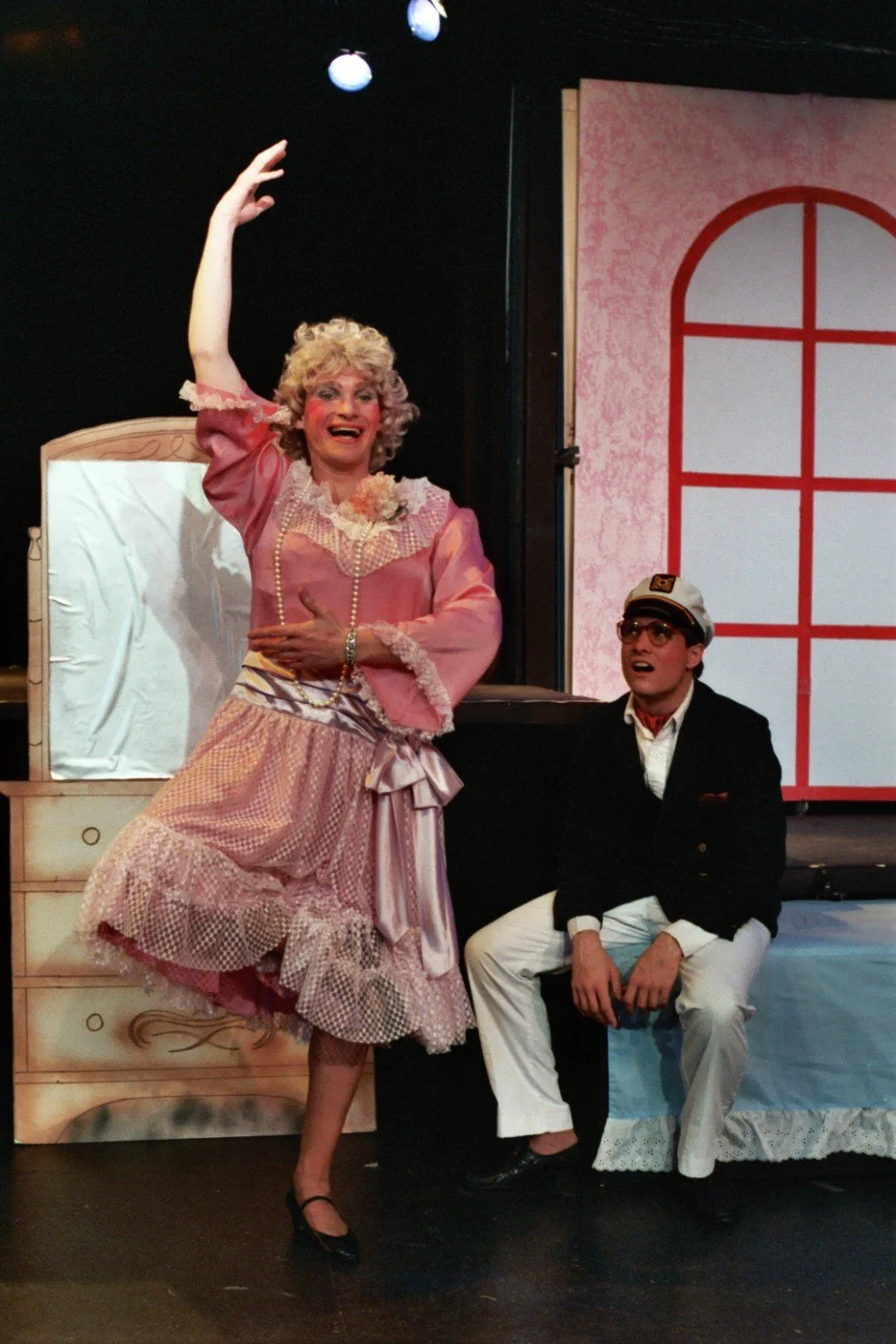 Two actors on stage in theatrical costumes. One is dressed in pink Victorian-style clothing, posing flamboyantly, and the other is sitting, dressed as a ship captain, wearing sunglasses and a hat.