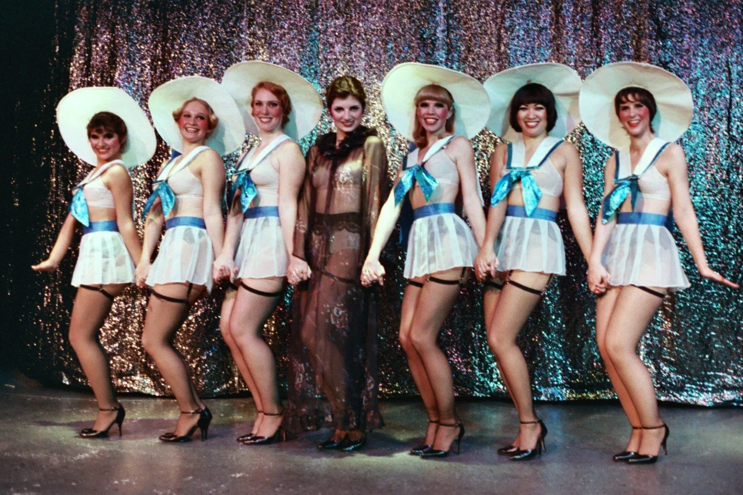 Group of performers in sailor-themed costumes with blue accents, large white hats, standing in front of a glittering curtain.