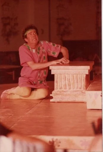 A person in a colorful costume is kneeling on stage, performing in a play, with hands on a small rectangular pillar. The stage is dimly lit, suggesting an indoor theater setting.