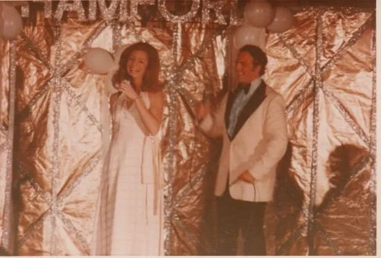 A woman and a man on stage with a metallic backdrop, holding microphones, dressed in formal attire.