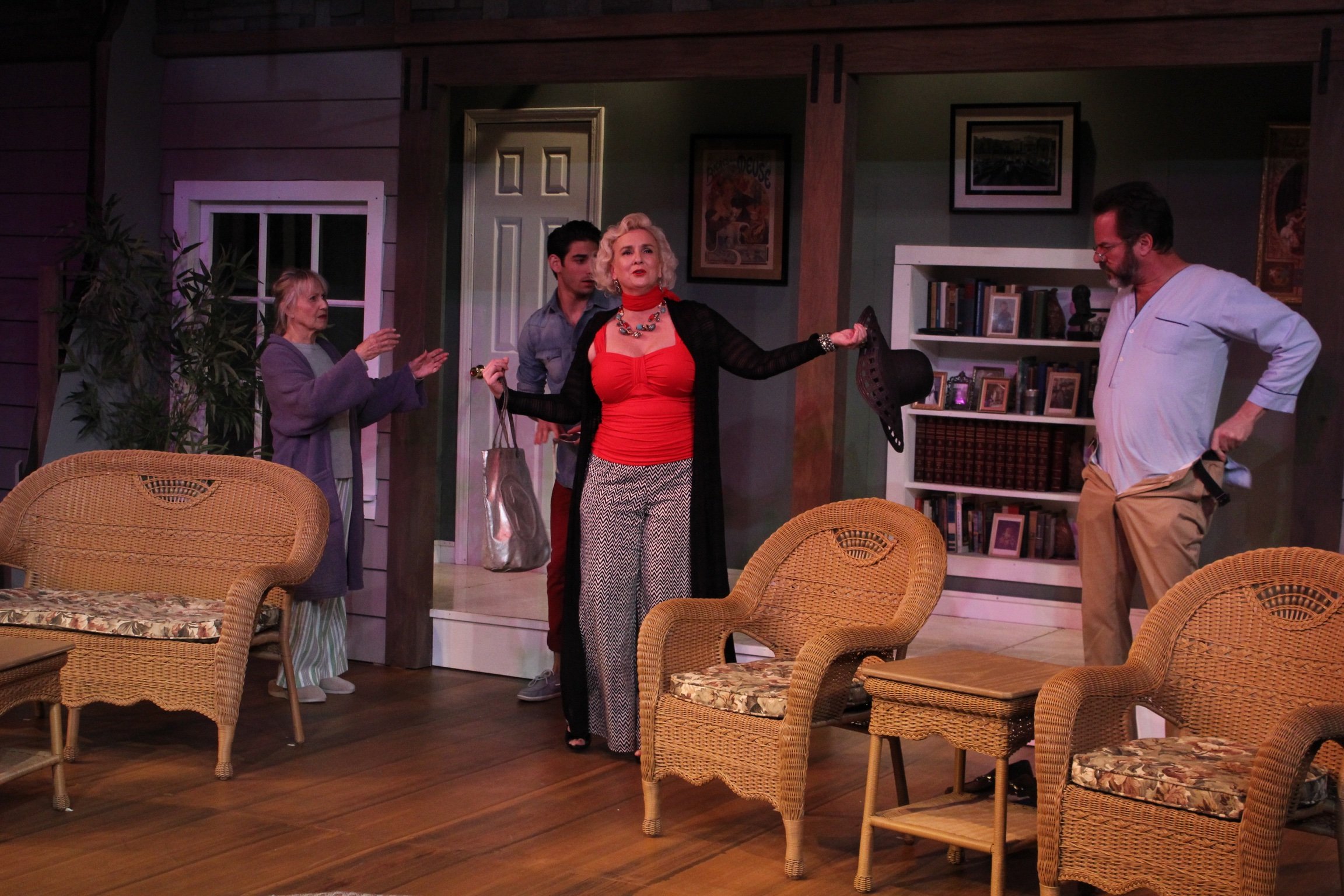 A theatrical performance featuring four actors in a room resembling a living space with wicker furniture. One actor, in a red top and black overlay, holds a hat; another wearing pajamas gestures towards her. A man adjusts his pants while another acto