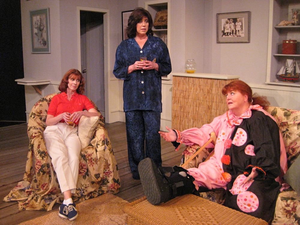 Three women in a living room setting, one sitting casually on a floral-covered chair, one standing in blue pajamas holding a drink, and the third dressed in a colorful clown costume with a crutch, sitting on a couch.