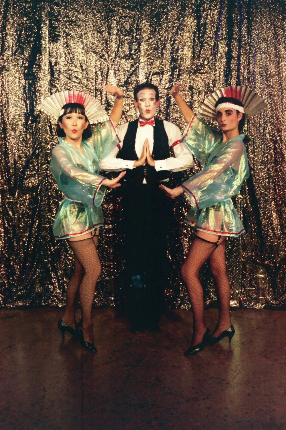 Three performers in theatrical costumes, two women in metallic dresses and headdresses flanking a man in mime makeup and formal attire, posing in front of a glittery backdrop.