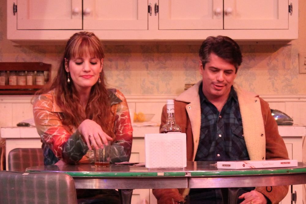 Two people sitting at a kitchen table with drinks, a bottle, and a napkin holder. The kitchen is styled with vintage cabinets and decor.