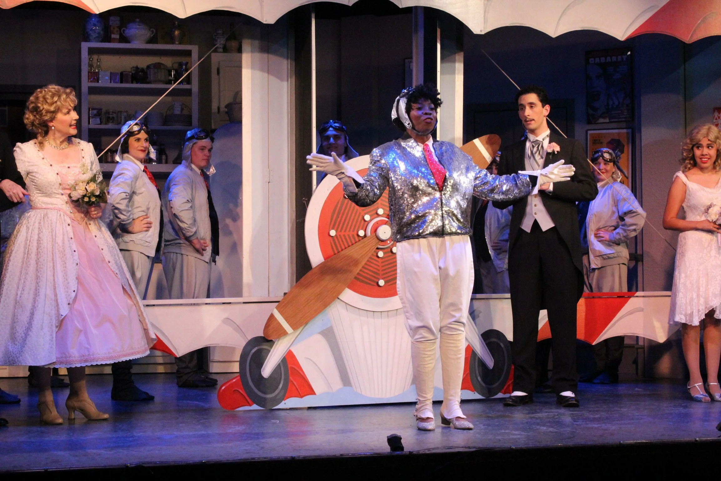 Stage performance with actors in vintage costumes, featuring a performer in a sparkly jacket, surrounded by people dressed in formal attire and aviator outfits near a propeller set piece.