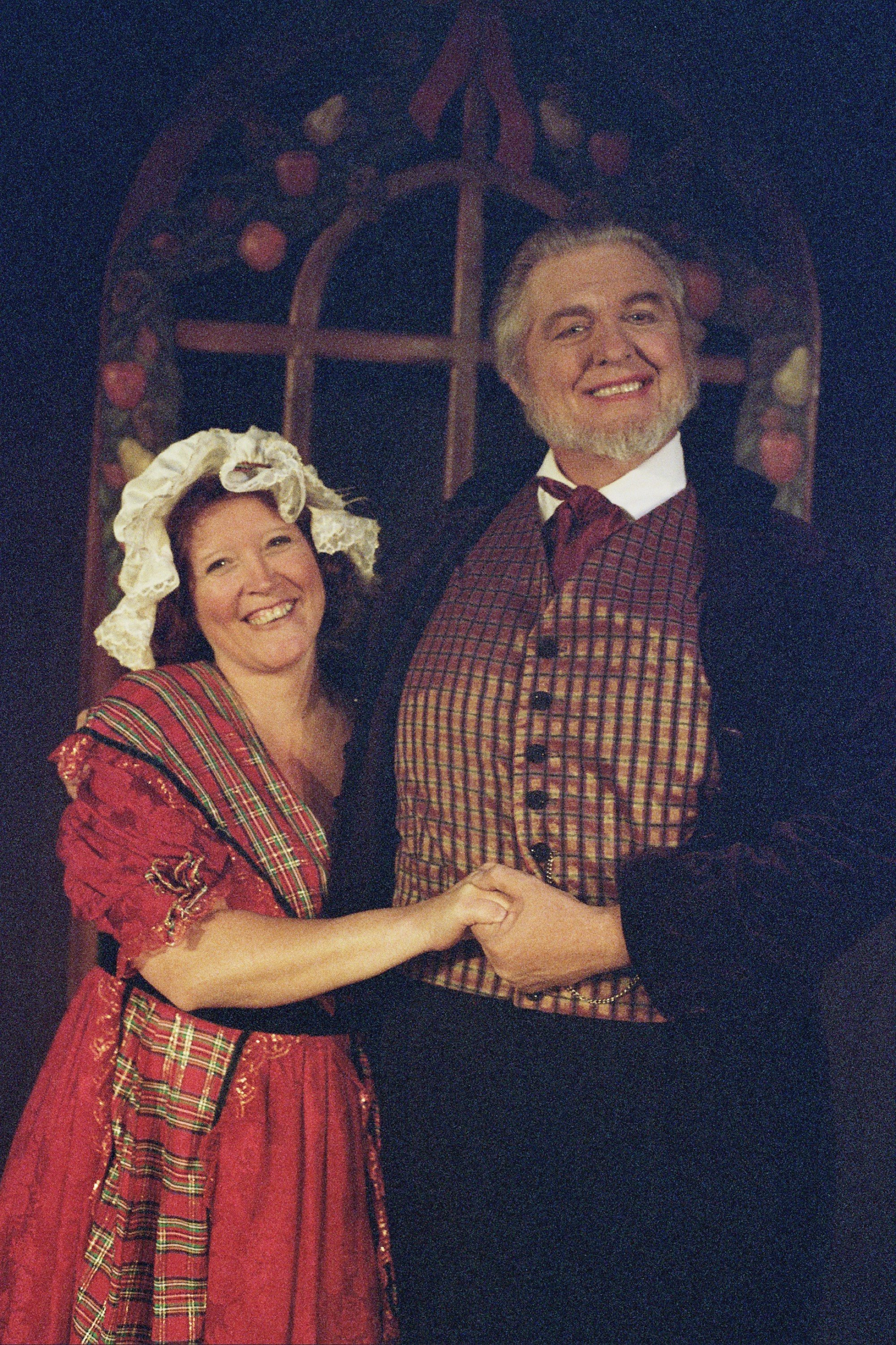 Two people in period costumes, a woman in a red dress and bonnet, and a man in a checkered vest and dark jacket, smiling and holding hands in front of a decorative backdrop.