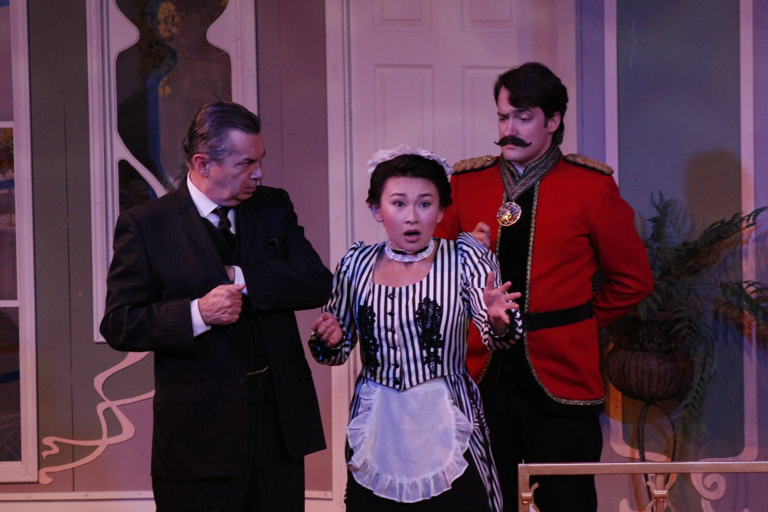 Three actors in theatrical costumes performing a dramatic scene inside a room, featuring a serious man in a suit, a surprised woman dressed as a maid, and a man in a red military uniform with a mustache.