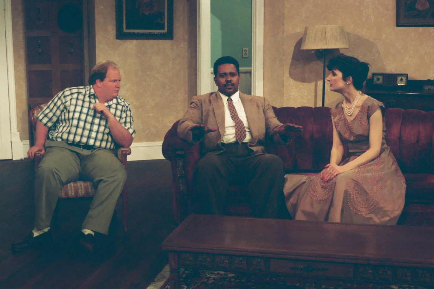 Three people sitting on a vintage couch in a living room set, dressed in mid-20th century attire; the man on the left is in a plaid shirt, the man in the center is in a suit, and the woman on the right is wearing a dress and pearls.