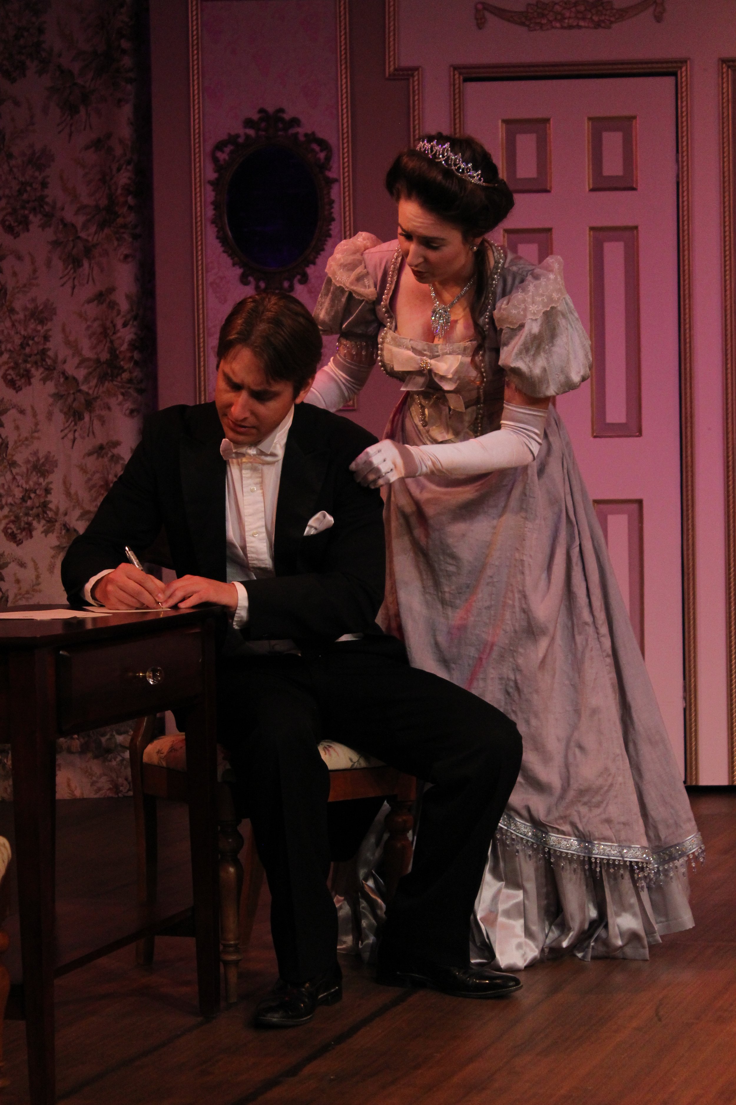 Victorian-era scene with a man writing at a desk and a woman standing beside him, set in an ornate room.