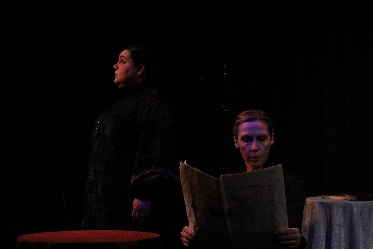 Two people on stage during a dramatic performance, one standing and looking up, the other sitting and reading a newspaper in a dimly lit setting.