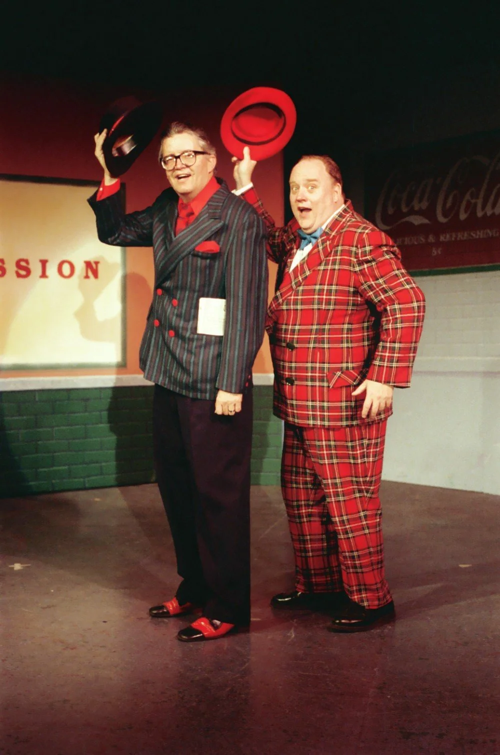 Two men in colorful suits holding hats, standing on a stage with a vintage Coca-Cola sign in the background.