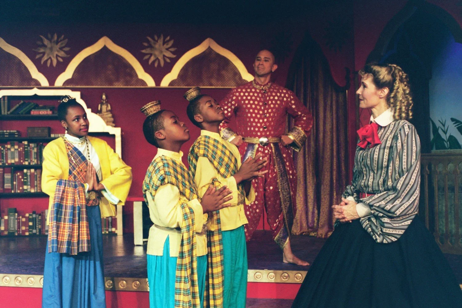 Stage performance with a group of actors in traditional Asian-inspired costumes, featuring a woman in a Victorian-style dress talking to two young boys in yellow attire and a man in a red outfit in the background, set against a detailed backdrop.