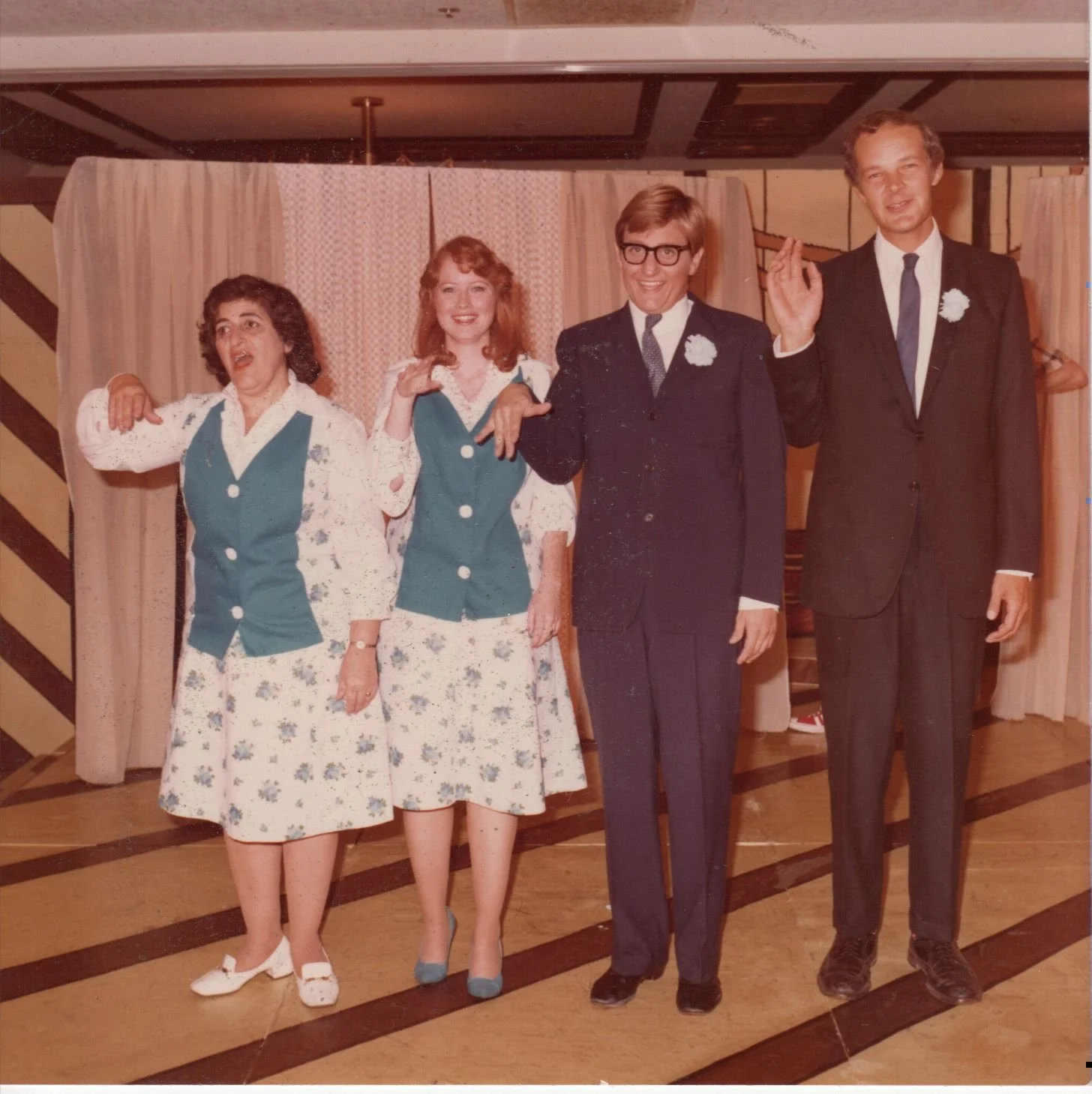 A vintage photo featuring two women and two men posing indoors. The women wear matching floral skirts and teal vests, while the men are in dark suits. They stand in front of curtains and appear to be mimicking hand gestures.