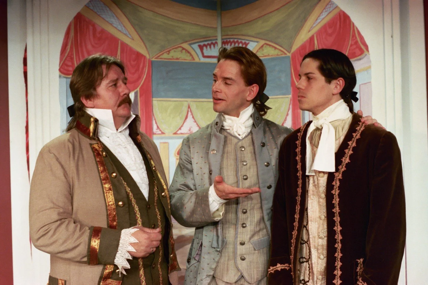 Three men dressed in 18th-century period costumes, wearing waistcoats and cravats, in front of a painted theatrical backdrop.
