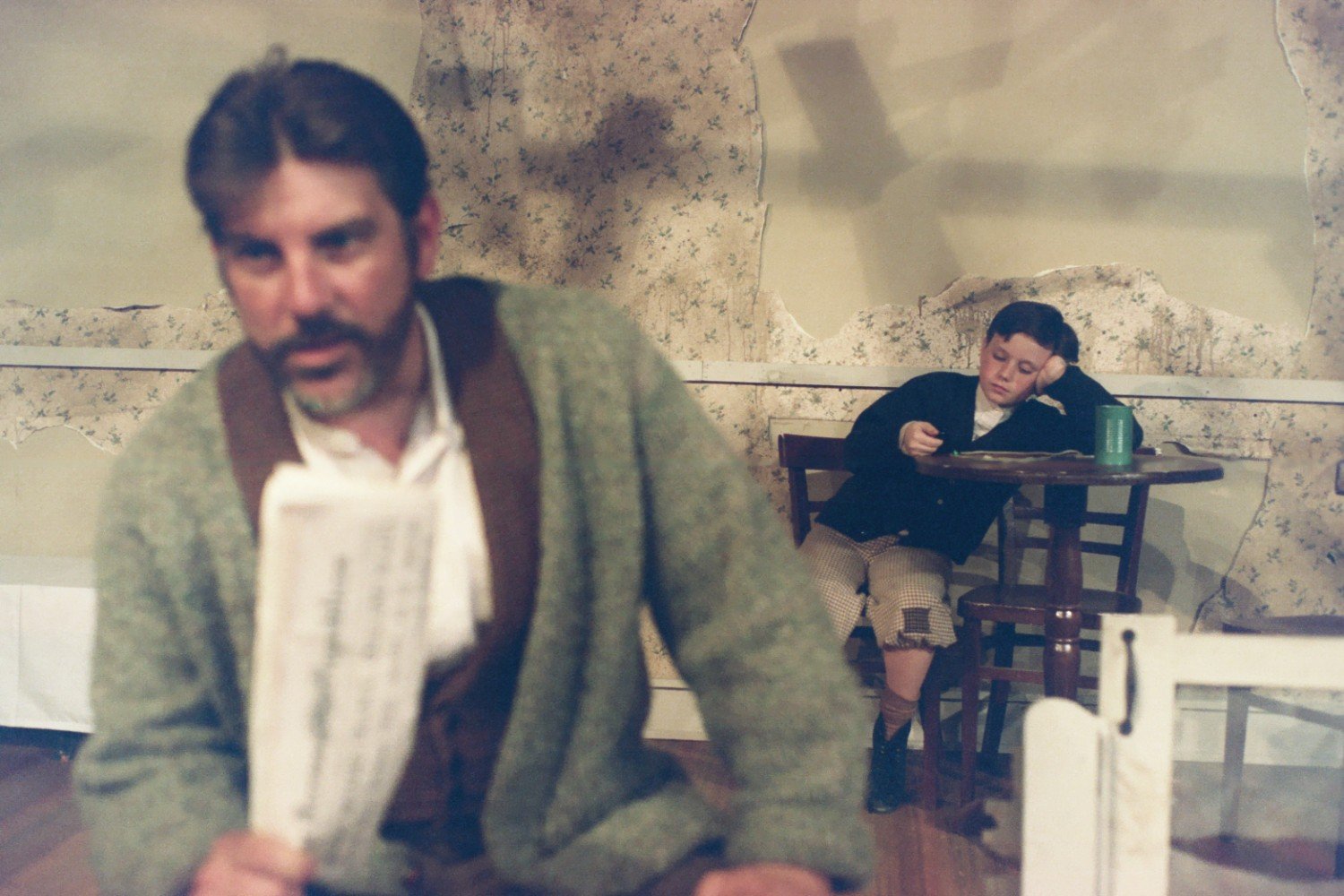 A blurred man in the foreground holding a piece of paper with a child sitting at a table in the background, appearing bored or tired. The setting has a rustic, old-fashioned feel with a worn wall and simple furniture.