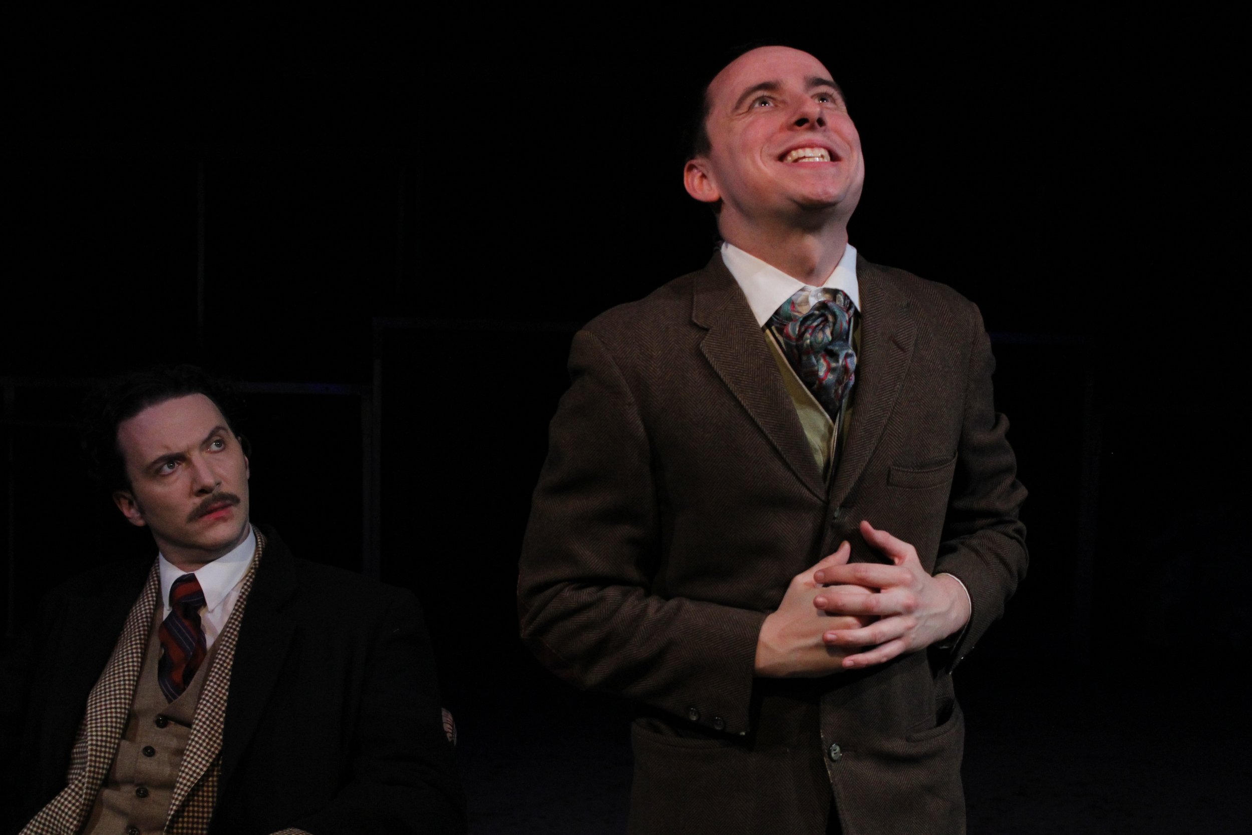 Two men in 1940s-style suits on a dark stage, one standing and smiling up, the other sitting and looking at him with a skeptical expression.