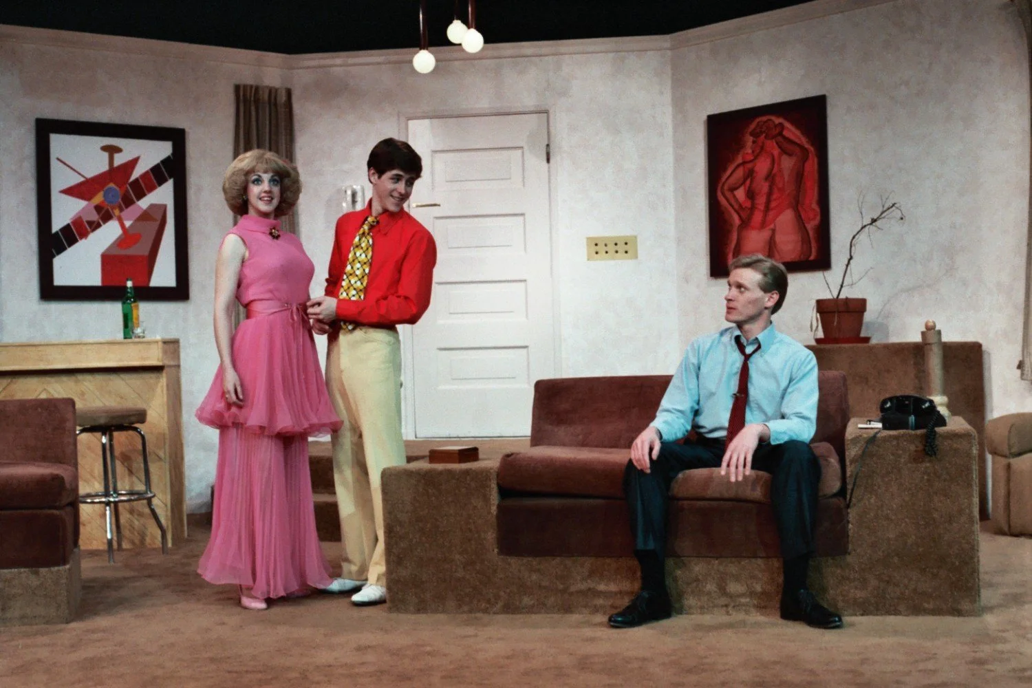 A theater stage depicting a living room scene with three actors. One actor is sitting on a sofa wearing a blue shirt and tie. Another actor, in a pink dress, and a third actor in a red shirt and yellow tie, stand together. The set includes abstract a