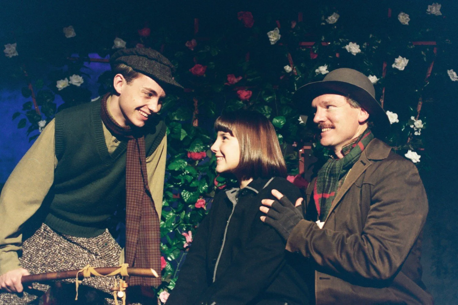 Three people in vintage clothing smiling in front of a background with roses and foliage.