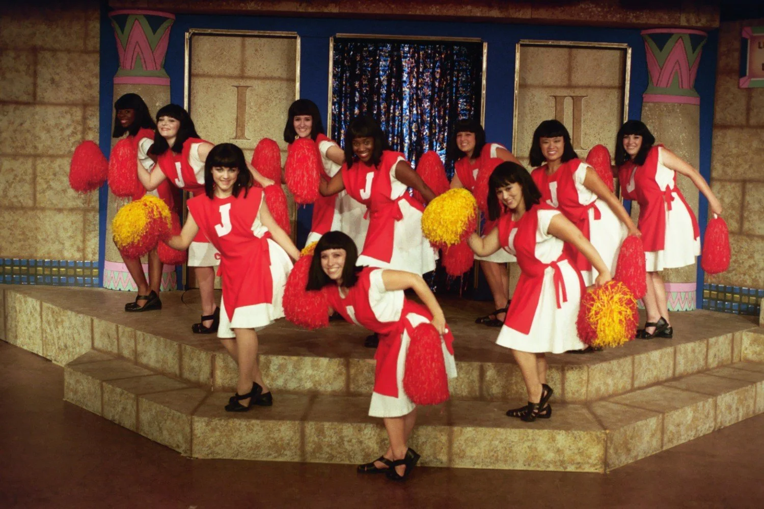 Group of cheerleaders in red and white costumes with letter 'J' and holding red and yellow pom-poms on a stage.