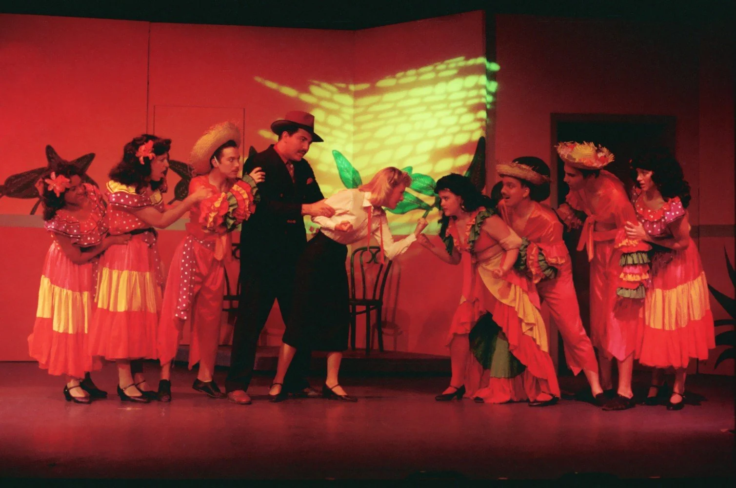 A theatrical performance with actors in colorful costumes on stage, engaging in a dramatic scene with vibrant red lighting.