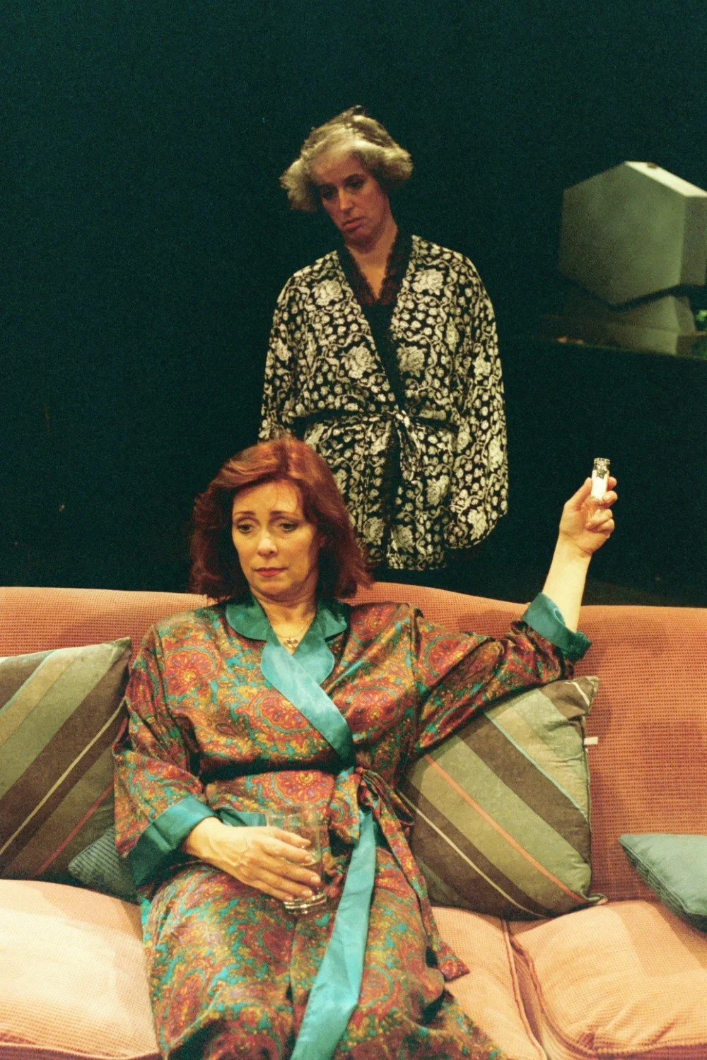 Two people in robes; one sitting on a couch holding a glass, another standing behind.