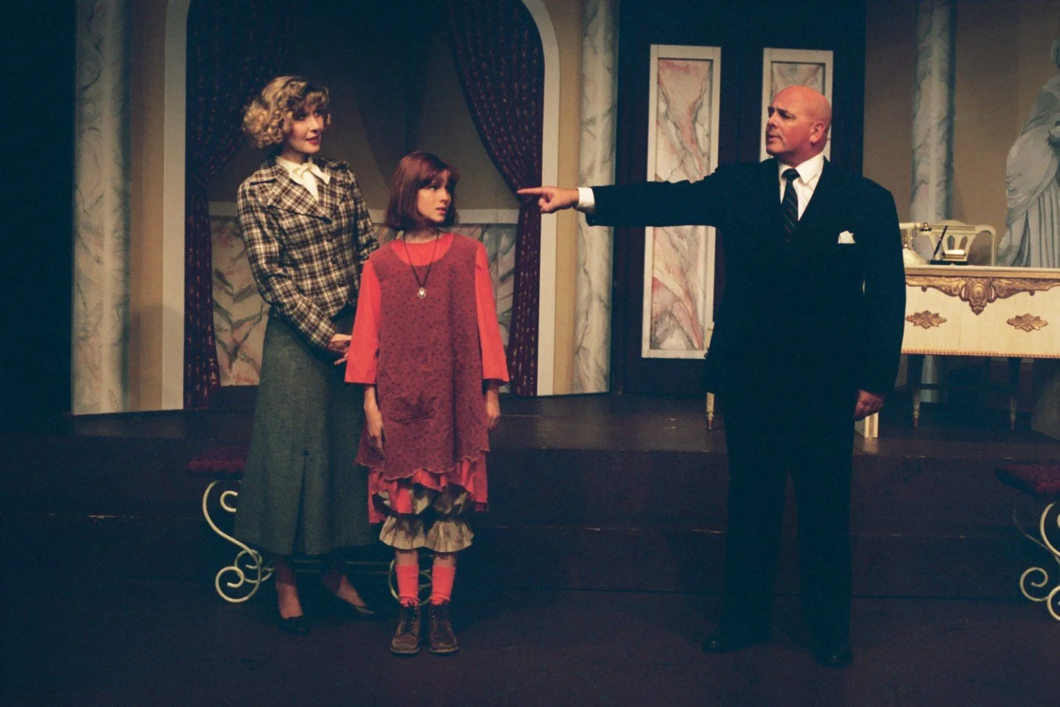 Three actors performing on a theater stage, with one pointing dramatically, while another stands with a child in period clothing. The set includes a desk and decorative columns.