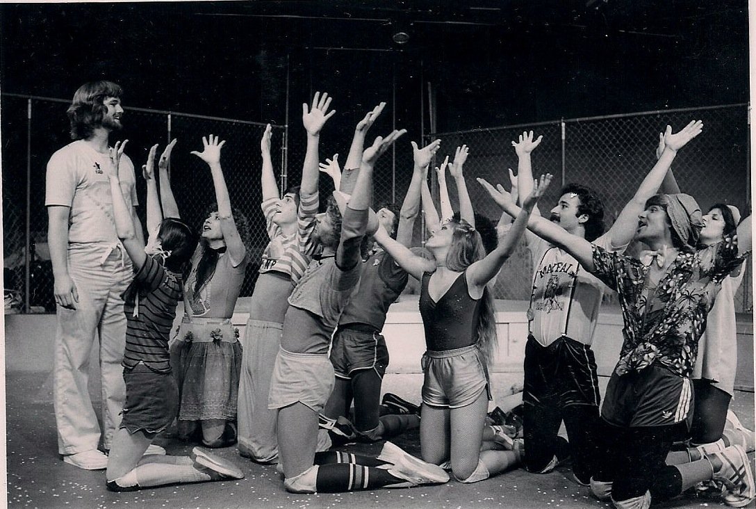 A group of people, mostly kneeling, with arms raised in a theatrical pose, dressed in casual and theatrical attire, possibly rehearsing or performing a scene, inside a fenced area.