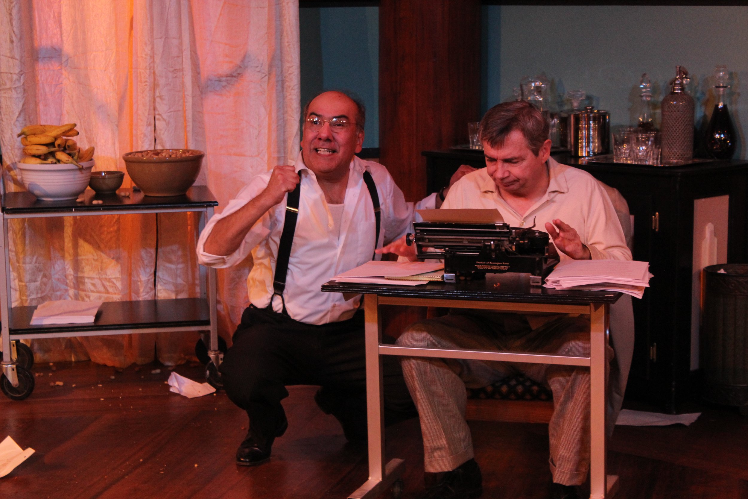 Two men are in an indoor setting with one sitting at a desk with a typewriter and papers. The other man, wearing suspenders and a white shirt, appears animated and is kneeling next to him. A cart with a bowl of bananas and a wooden bowl is in the bac