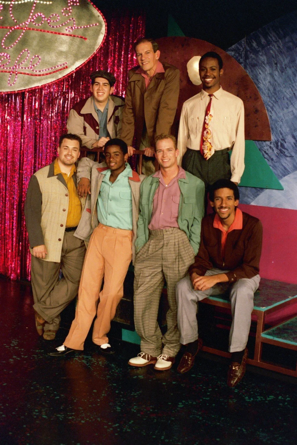 A group of seven men in vintage-style attire, posing on a stage set with colorful background and a sign that says "Smokey Joe's Cafe." The men are smiling, with some seated and others standing, against a backdrop of shiny red curtains and geometric s
