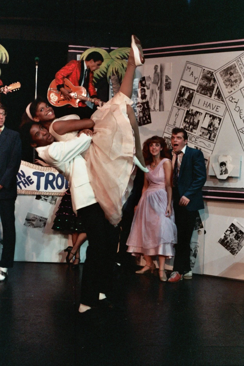A lively scene of a dance performance with a man lifting a woman in the foreground, musicians playing guitar in the background, and other people watching. The setting is festive, with decorations and a backdrop displaying photos and graphics. The wom