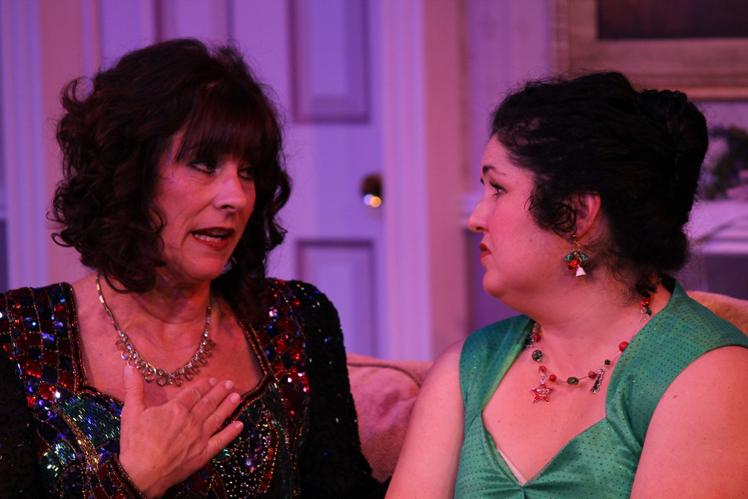 Two women sit close together in conversation, one wearing a colorful sequined dress and necklace, the other in a green dress with festive jewelry. They appear engaged in an emotional exchange.