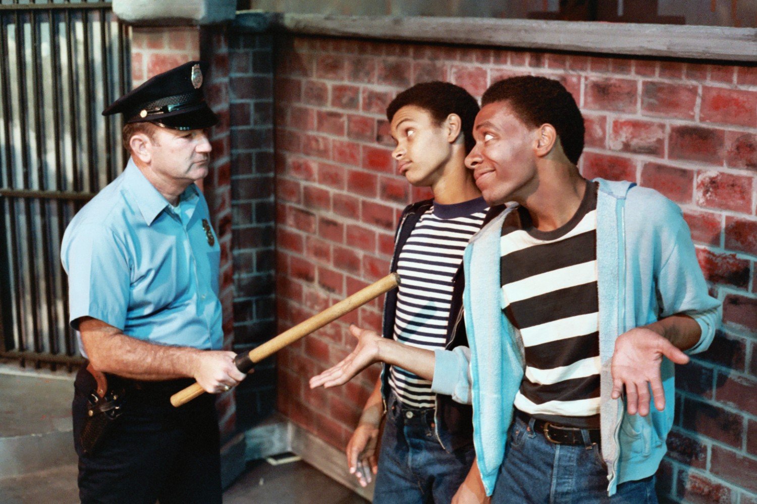 A police officer holding a baton confronts two young men in striped shirts standing against a brick wall. The young men appear to be in a casual or dismissive posture.