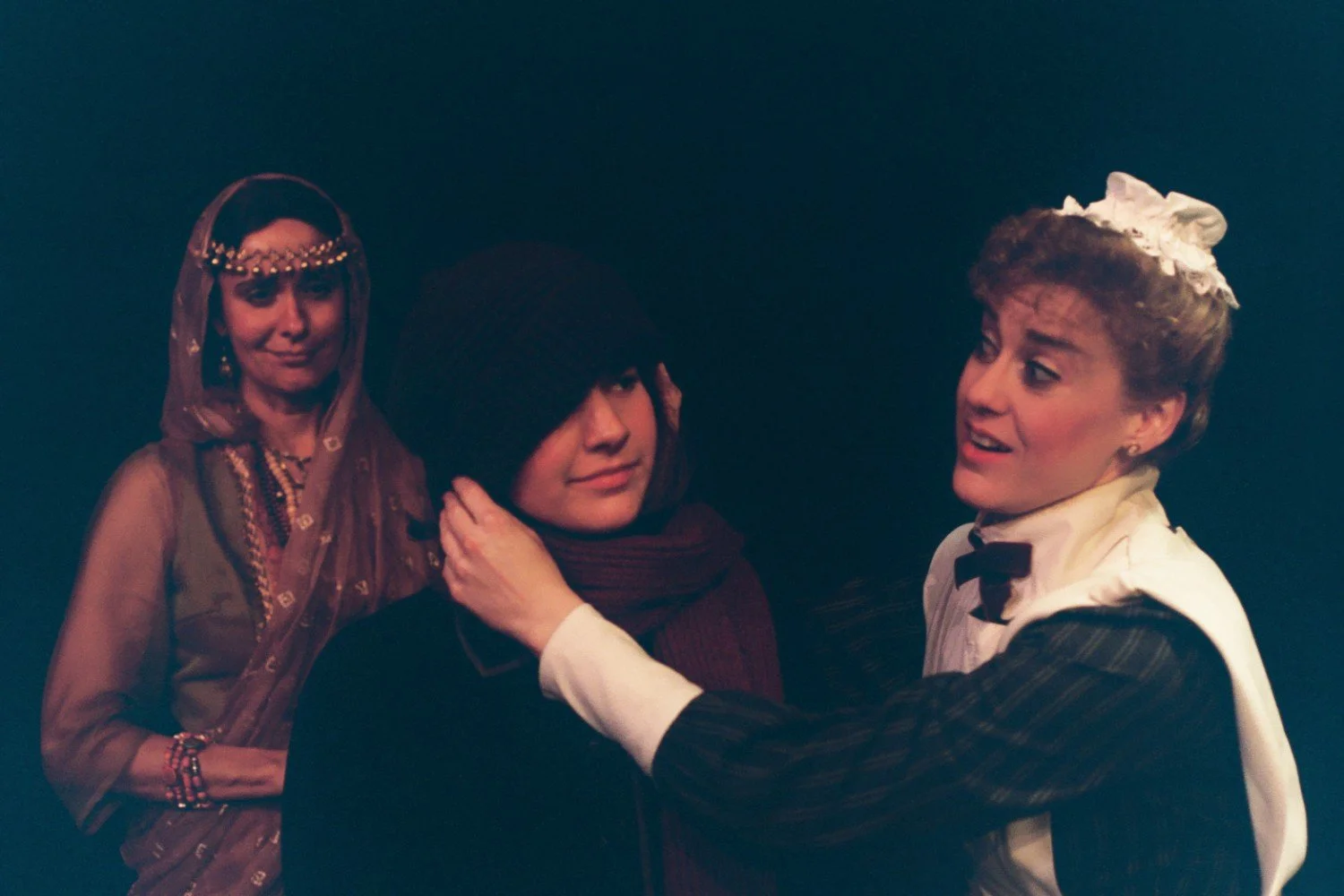 Three actors in period costumes; one in a sari with headdress, another in dark winter attire, and the third in a maid costume adjusting a scarf.