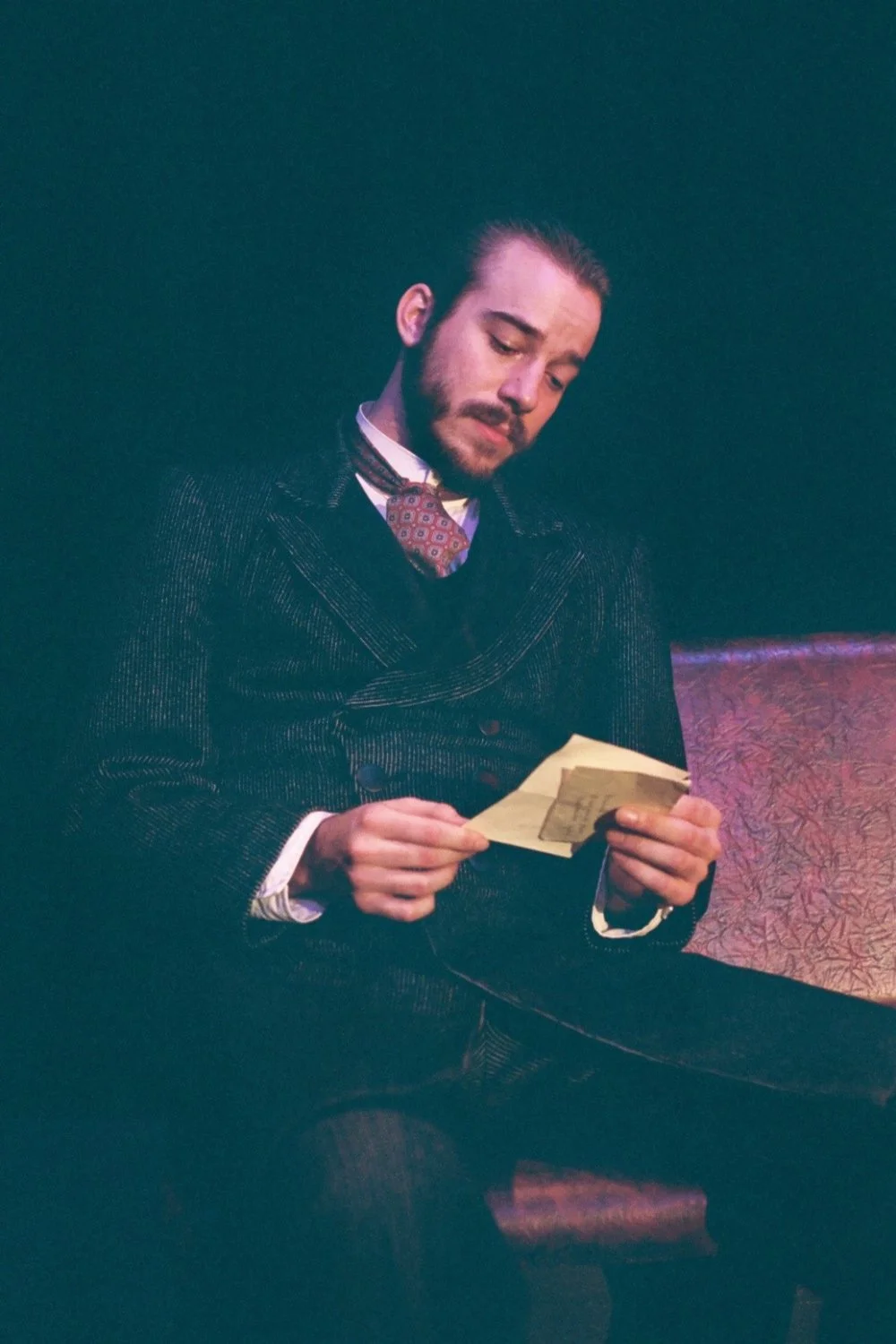 A man in a vintage suit sitting, reading a paper note in a dimly lit room.