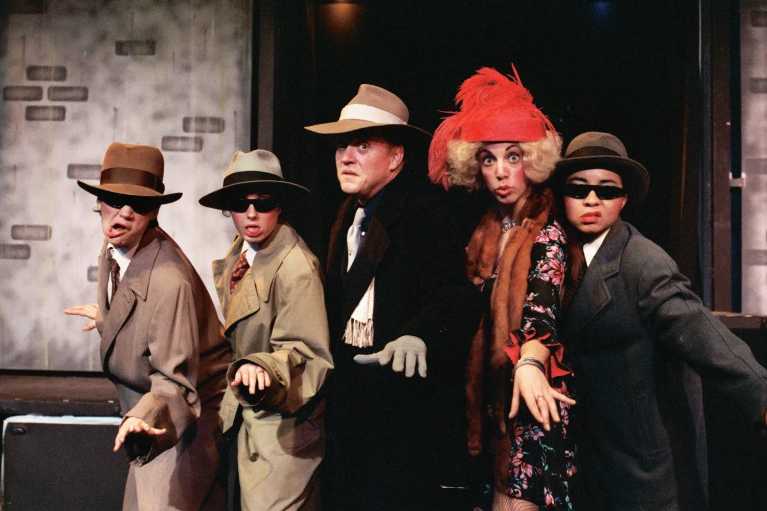 Five people in theatrical costumes resembling film noir characters, with exaggerated expressions and poses, wearing trench coats, fedoras, sunglasses, and a flamboyant red hat.