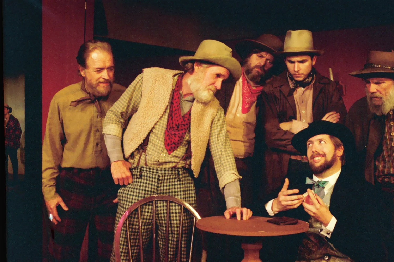Group of men in Western attire gathered around a table in a theatrical setting.