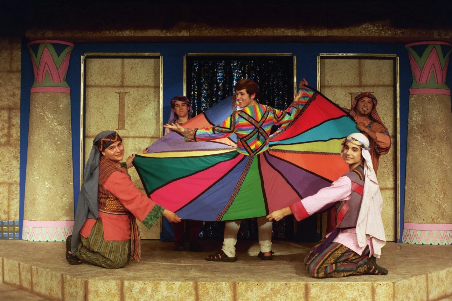 A stage performance featuring a person wearing a colorful, striped coat, surrounded by three others in traditional-style clothing. The setting resembles an ancient Egyptian or Middle Eastern backdrop with painted columns.