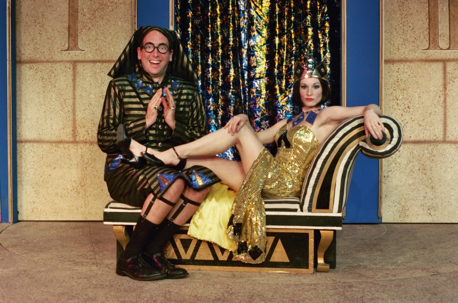 Two people in Egyptian-themed costumes, one sitting on a chaise lounge in a gold sequin dress with a headdress, the other kneeling with a gleeful expression, wearing a striped outfit and glasses.