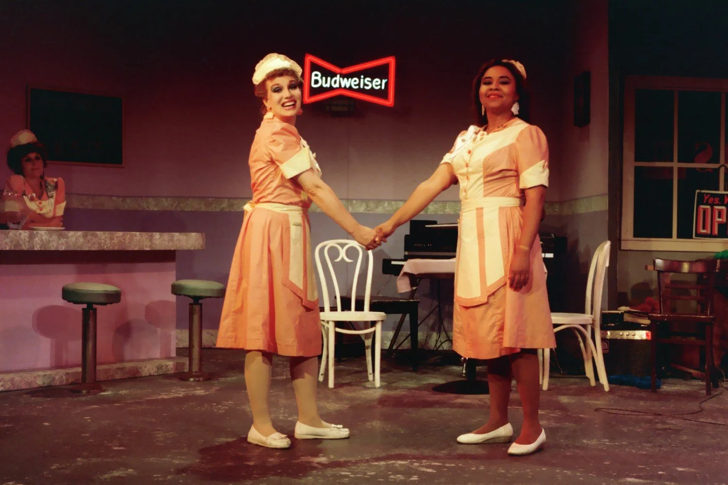 Two women in diner uniforms holding hands on stage, with a Budweiser sign in the background and a woman sitting at a counter.