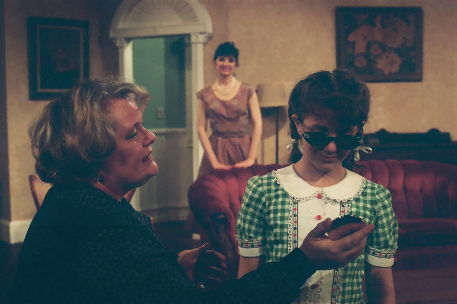 Two women and a girl in a living room setting, with the girl wearing sunglasses and a green checkered dress. A woman in the foreground shows something to the girl, while another woman stands in the background, smiling.