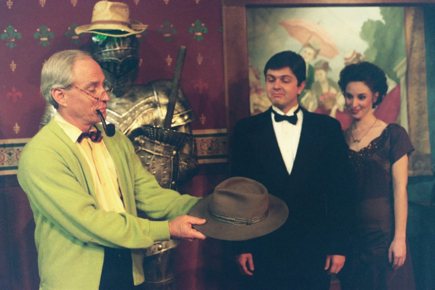 A man in a green cardigan holding a hat and smoking a pipe, stands near a knight's armor. Next to him, a couple dressed in formal attire, a man in a tuxedo and a woman in a brown dress, look on with curiosity. The background features a colorful paint